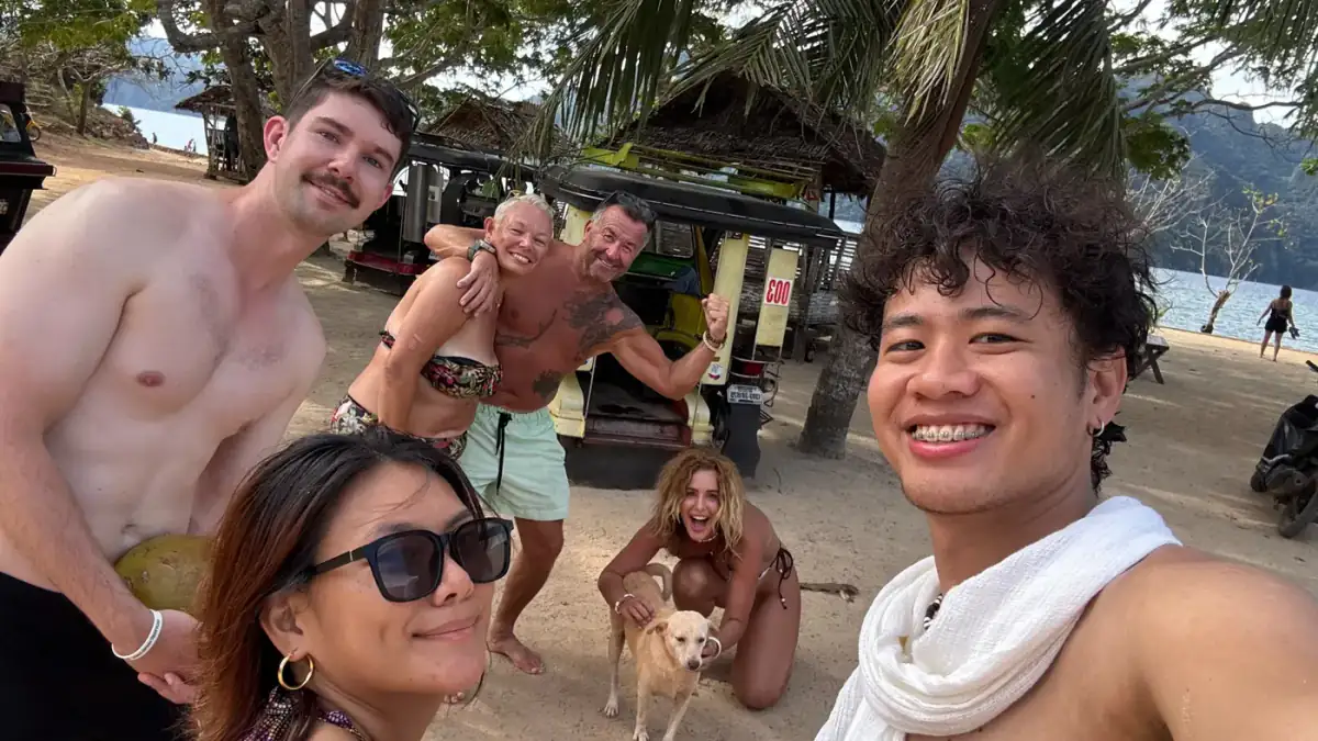Guests laughing on the beach with a dog, a tuk-tuk behind them